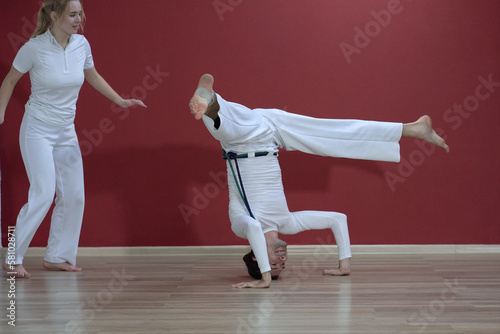 A mature man, a sports master, and a young woman, an aspiring athlete, practice the Brazilian martial art of capoeira in a small gym.