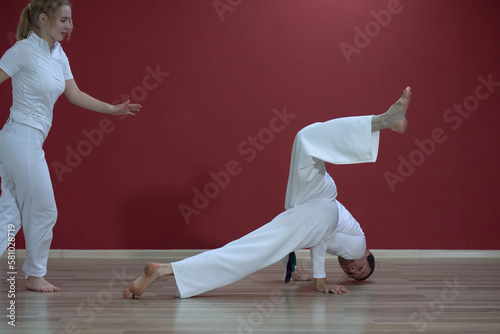 A mature man, a sports master, and a young woman, an aspiring athlete, practice the Brazilian martial art of capoeira in a small gym.