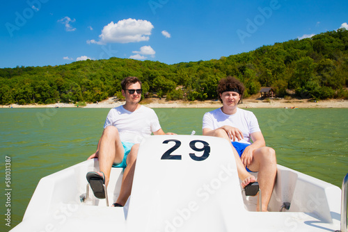 Two young men on water pedal boat tourist catamaran on mountain lake on sunny day. Summer leisure activities in public park. European caucasian white young male men. Lifestyle portrait