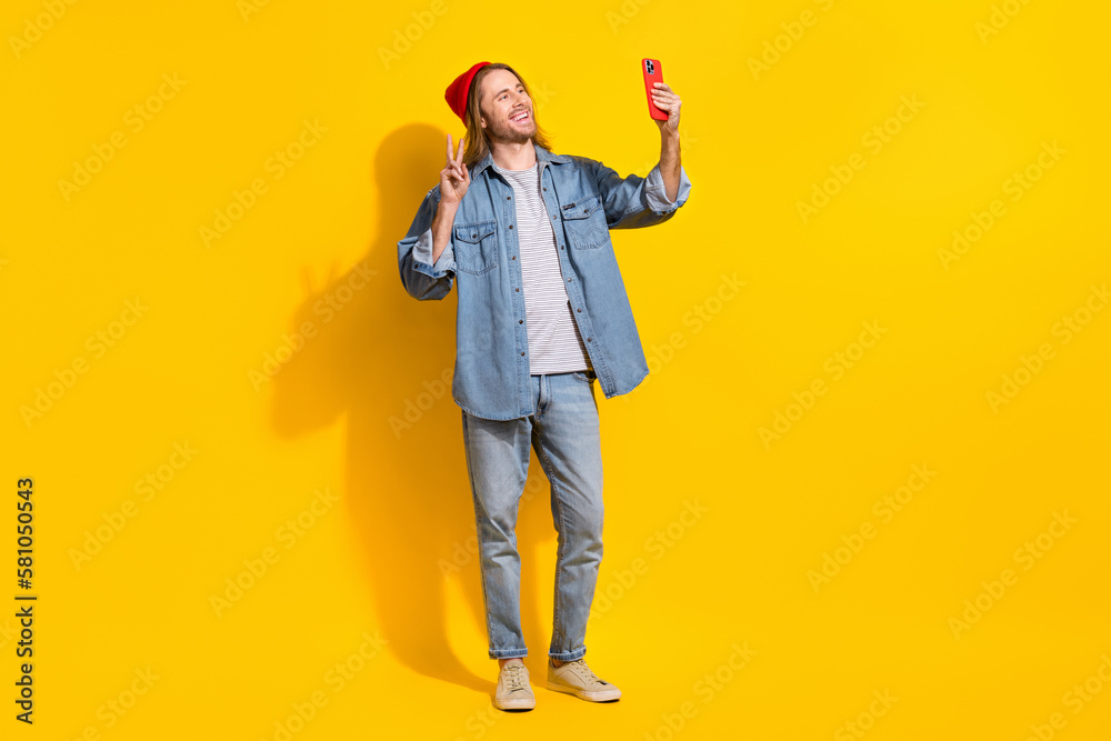 Photo of cheerful satisfied optimistic guy long hairstyle jeans shirt making selfie on front camera isolated on yellow color background