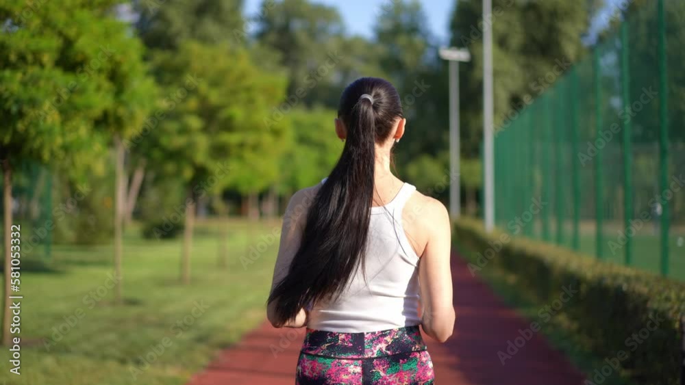 Back view slim woman with long ponytail jogging in slow motion on sunny day in park. Tracking shot of confident fit Caucasian sportswoman running on sports track outdoors Back view slim woman with long ponytail jogging in slow motion on sunny day in park. Tracking shot of confident fit Caucasian sportswoman running on sports track outdoors