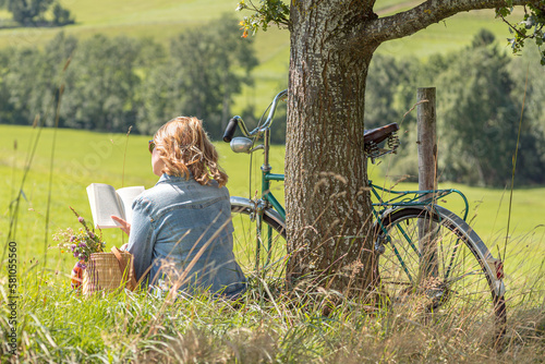 Relaxed blonde Woman reading book under tree. Retro bicycle and basket with flowers.