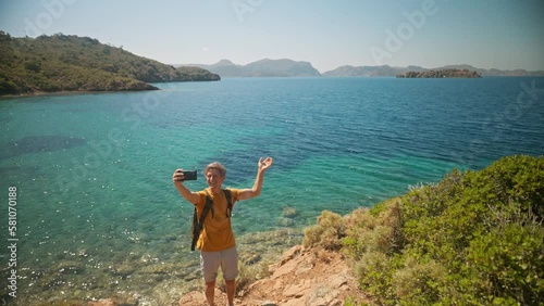 happy free man traveler with backpack makes selfie or video call standing on cliff on shore of beautiful sea bay with clear turquoise water and paradise empty sea beach. slow motion, travel in Turkey photo