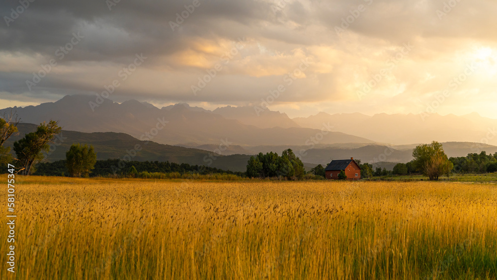 The Sun shines through the clouds after an afternoon rain in the San Juan Mountains, southwestern Colorado. Sunlight falls on the landscape below, lighting up a golden wheat field and a distant barn.