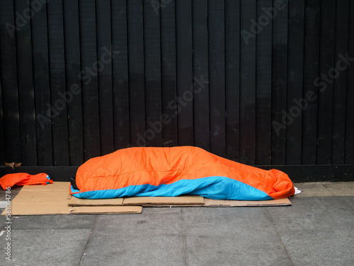 A homeless rough sleeper on the pavement in Central London. Only an orange sleeping bag and cardboard box flooring to keep dry and warm. Concept for poverty, economy, social decline, on the streets.