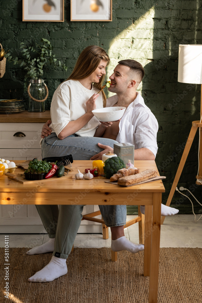 Young happy couple in the kitchen. A smiling woman feeds her husband sitting on his lap. Full length photo. Young happy couple in the kitchen. A smiling woman feeds her husband sitting on his lap. Full length photo.
