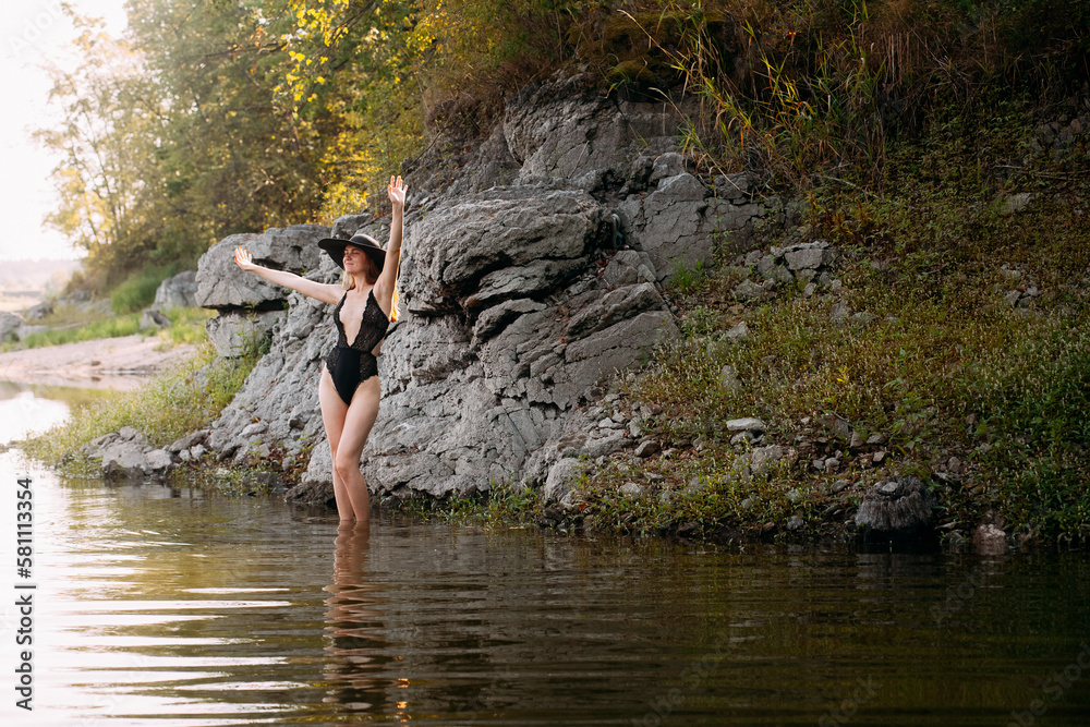 Fototapeta premium A young girl with long blond hair and a beautiful figure, in black openwork lingerie and a black wide hat, stands against the backdrop of a rock, on the banks of a picturesque river.