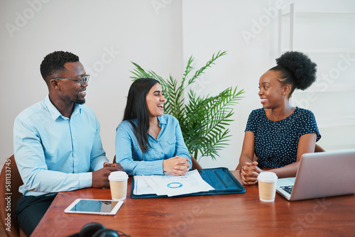 Three colleagues laugh during informal meeting at boardroom table with coffee