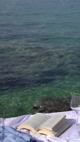 A vertical video of a book on a white beach blanket with a blue shirt and a glass of wine next to the sea