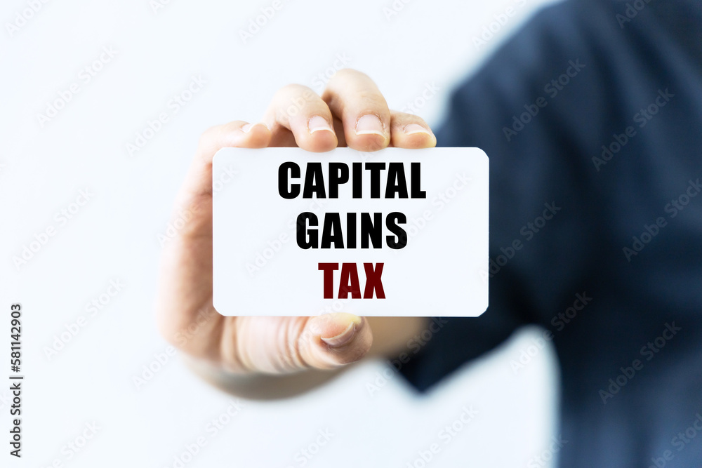 Capital gains tax text on blank business card being held by a woman's hand with blurred background. Business concept about capital gains tax. Capital gains tax text on blank business card being held by a woman's hand with blurred background. Business concept about capital gains tax.