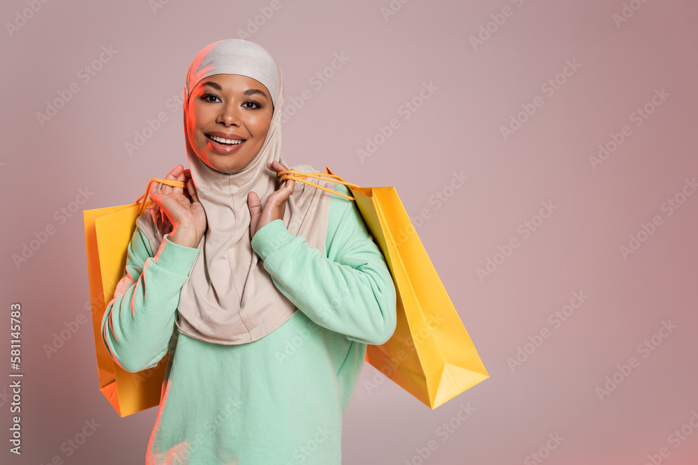 overjoyed multiracial woman in muslim hijab holding yellow shopping bags and smiling at camera on pinkish grey. overjoyed multiracial woman in muslim hijab holding yellow shopping bags and smiling at camera on pinkish grey.