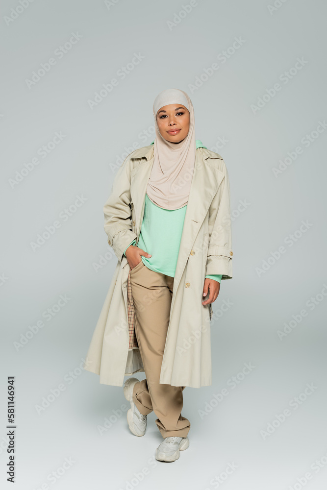 full length of multiracial woman in fashionable casual clothes and hijab standing with hand in pocket on grey background.