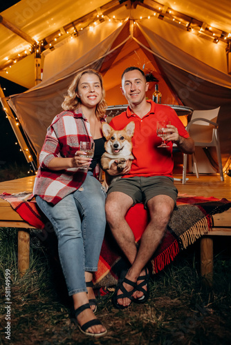 Happy lovely couple with Welsh Corgi Pembroke dog relaxing in glamping on evening and drinking wine near cozy bonfire. Luxury camping tent for outdoor recreation and recreation. Lifestyle concept