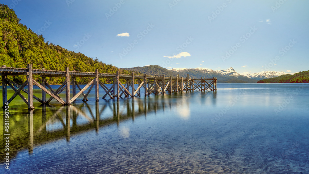 A beautiful pier to the left of the lake in shades of green and blue with a background of mountains in the morning.