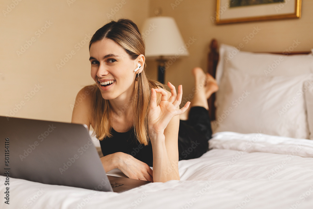 Happy woman in wireless earphones using laptop making video call lying on bed at home in bedroom and show all right. Modern communication. Freelancer lady browsing internet. Technology and freelance. Happy woman in wireless earphones using laptop making video call lying on bed at home in bedroom and show all right. Modern communication. Freelancer lady browsing internet. Technology and freelance.