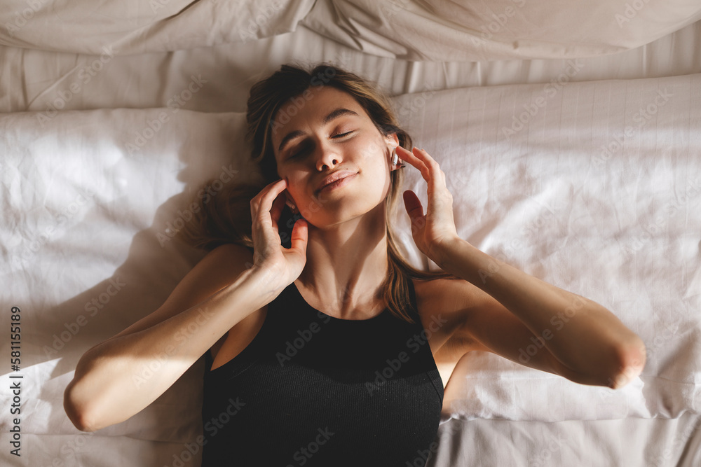 Naklejka premium Beautiful young woman listening music in wireless headphones at home lying on bed. Young pretty woman person are happy and enjoy with music by using earphone for listen a sound audio in bedroom.