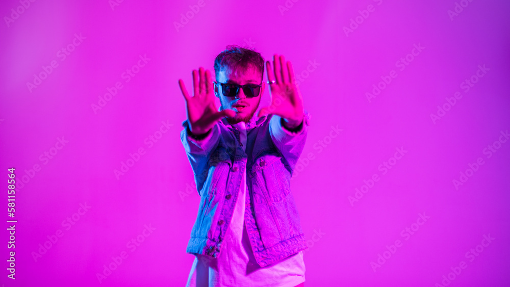 Handsome young hip hop dancer man with stylish sunglasses in fashion jeans jacket dancing in colorful studio with creative pink and blue light Handsome young hip hop dancer man with stylish sunglasses in fashion jeans jacket dancing in colorful studio with creative pink and blue light