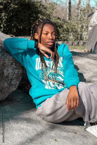 Mujer joven de color con trenzas en el pelo y vestida en chándal sentada en las piedras de un parque de Madrid con la mirada perdida pensativa. photo