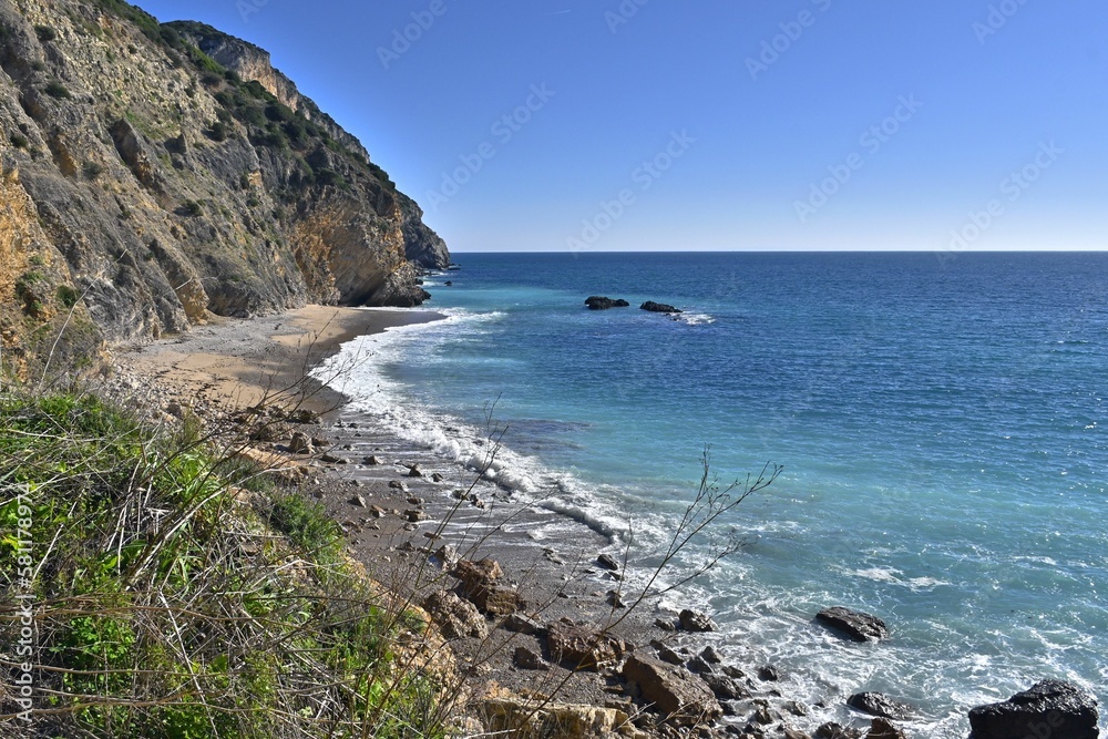View of Praia da Mijona Beach and blue sea surrounded by rocky hills under clear sky in Portugal