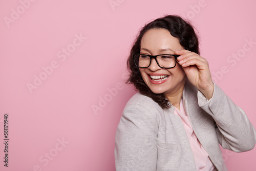 Portrait of a beautiful woman in stylish eyeglasses, smiling broadly looking aside, isolated on pink background with free ad space. Eyesight. Health. Medicine. Ophthalmology and fashion concept