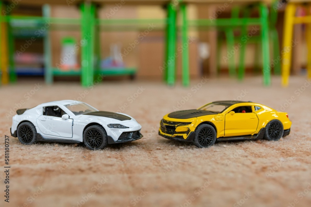 Two toy models of sports cars stand on piled brown carpet against abstract blurred background.