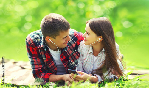 Portrait of happy smiling young couple listening to music in earphones with smartphone lying on the grass together in summer park