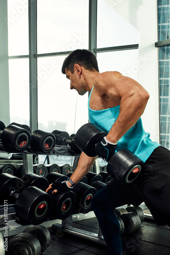 Man doing exercise with dumbbell leaning on sports bench in the gym. Photo of a sexy muscular man in sportswear and good physique on grey background. Strength and motivation, sport, fitness goal