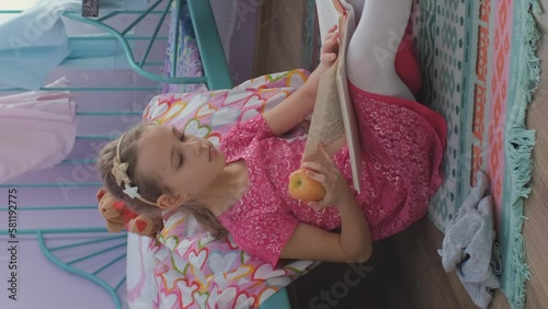 Vertical video. Little girl reading book and eating an apple in messy room. Literature and education concept photo