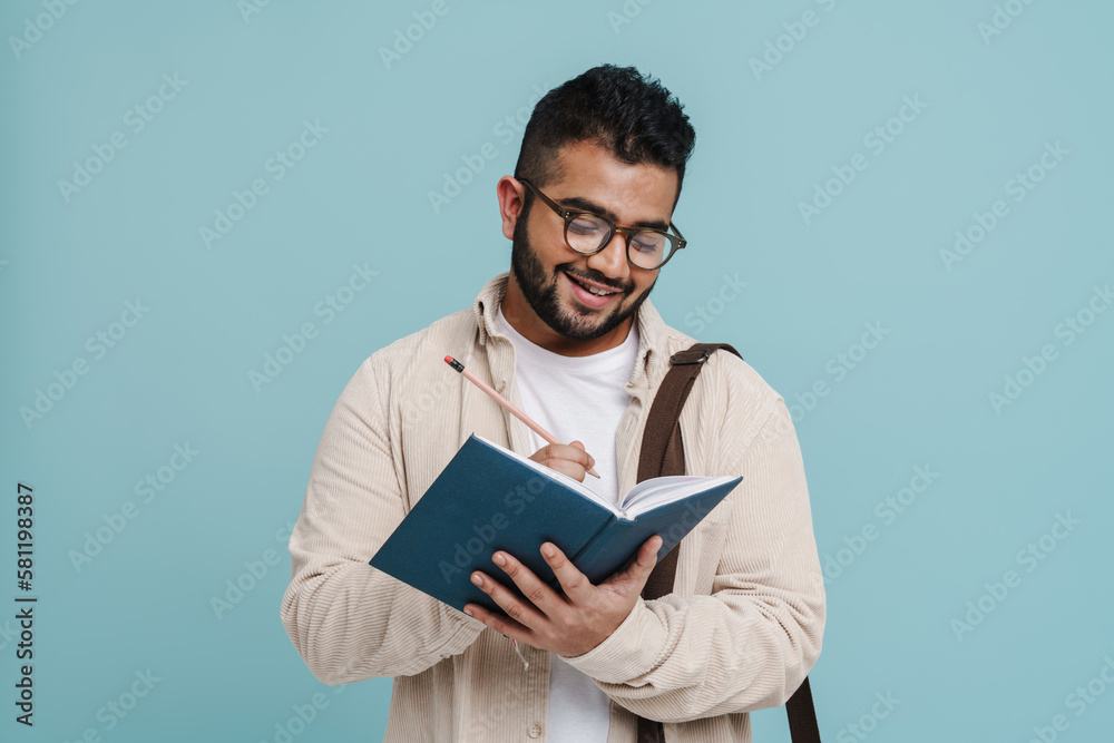 Smiling indian man writing down notes while standing isolated over blue background Smiling indian man writing down notes while standing isolated over blue background