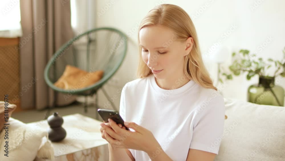 Young blonde woman is using phone swiping social media and smiling at home. Texting online with friends through a messenger or plays mobile games app. Young blonde woman is using phone swiping social media and smiling at home. Texting online with friends through a messenger or plays mobile games app.