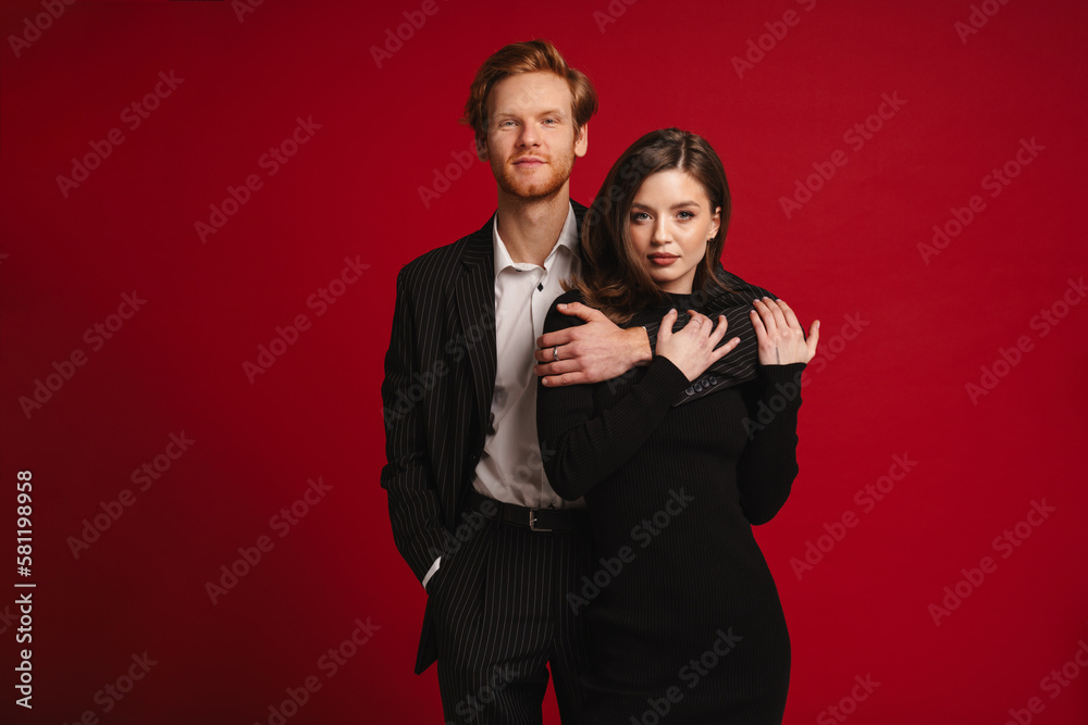 Beautiful couple hugging and looking at camera while standing isolated over red wall Beautiful couple hugging and looking at camera while standing isolated over red wall