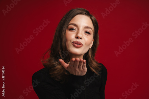 Woman blowing air kiss at camera while standing isolated over red background