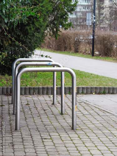 Empty bicycle stands on the pavement. Grass and trees in the background.