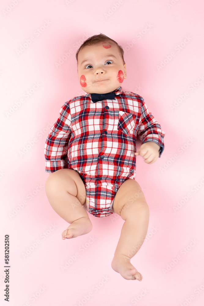 Child in beautiful plaid shirt on pink, traces of kisses with red lipstick on his face. Mockup for advertising, design, postcards, Valentine's Day. Child in beautiful plaid shirt on pink, traces of kisses with red lipstick on his face. Mockup for advertising, design, postcards, Valentine's Day.