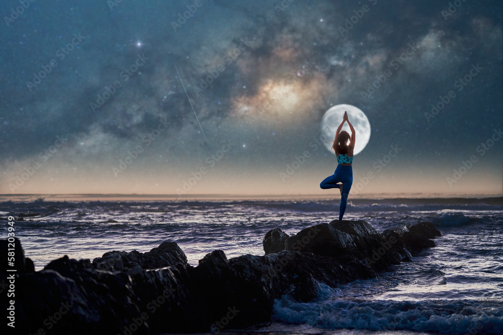 silhouette of a woman in yoga pose on rocks on the beach at night with the moon and milky way in the background	 - obrazy, fototapety, plakaty 