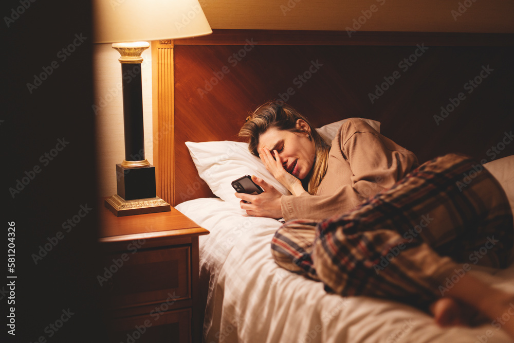 Young depressed woman using mobile phone crying on bed at night feeling sad and depressed victim of cyber bullying or broken heart suffering depression in desperate woman expression.