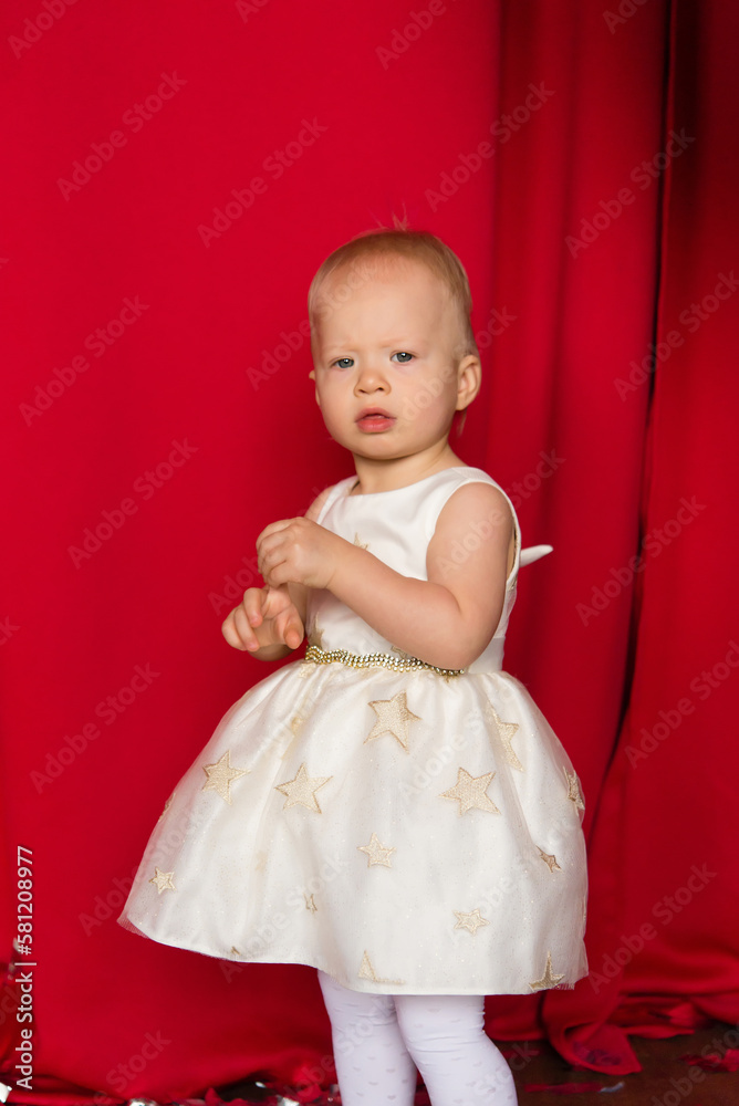 beautiful little toddler girl in a red dress against the background of red curtains. Birthday, new year, merry christmas beautiful little toddler girl in a red dress against the background of red curtains. Birthday, new year, merry christmas