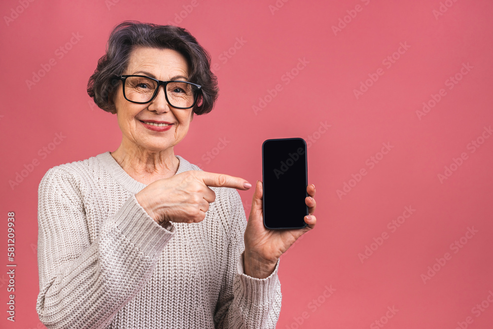 Surprised smiling happy senior mature aged senior woman in casual showing blank smartphone screen while looking at the camera isolated over pink background. Using phone. Surprised smiling happy senior mature aged senior woman in casual showing blank smartphone screen while looking at the camera isolated over pink background. Using phone.