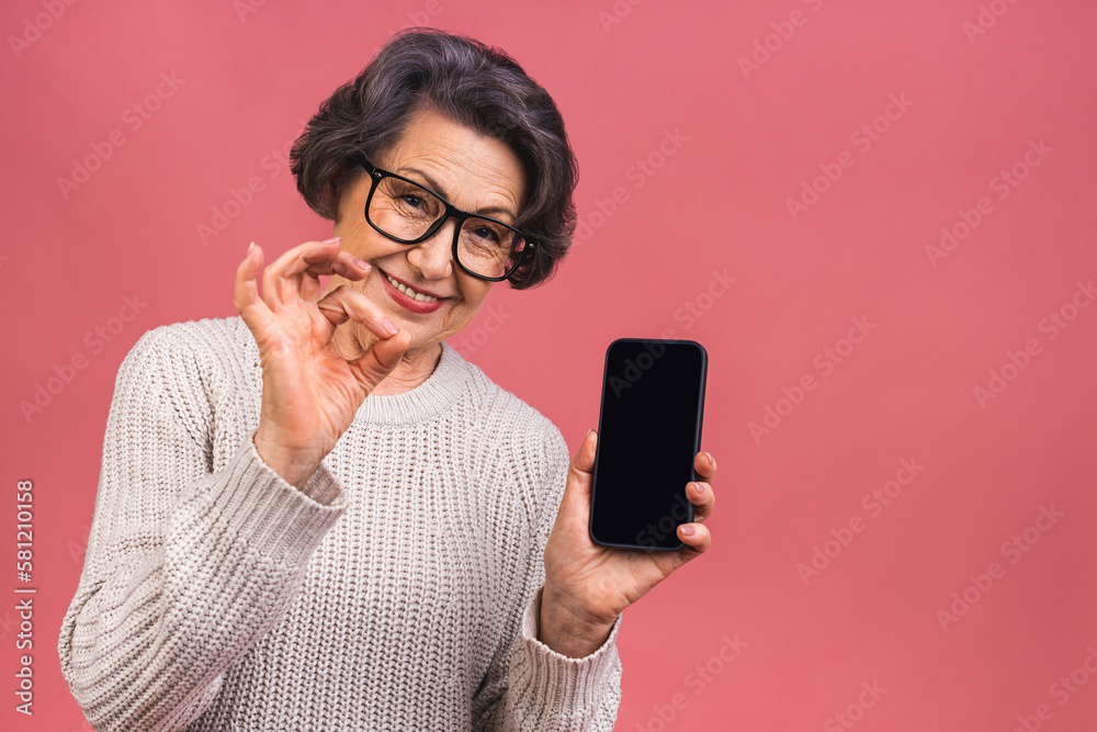 Surprised smiling happy senior mature aged senior woman in casual showing blank smartphone screen while looking at the camera isolated over pink background. Using phone. Ok sign.