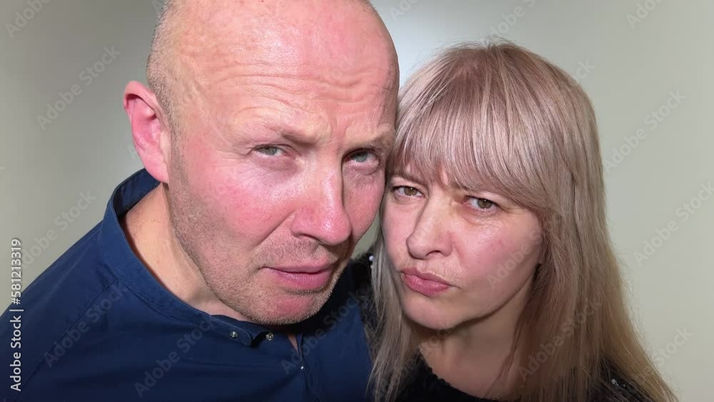 An adult couple looks with interest into the camera directly into the frame at us, they look as if asking who is there bald man in woman has blond hair on a white background in dark blue clothes An adult couple looks with interest into the camera directly into the frame at us, they look as if asking who is there bald man in woman has blond hair on a white background in dark blue clothes