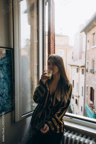 young beautiful woman in a black striped blouse drinks morning coffee by the window. morning ritual before the start of the day.