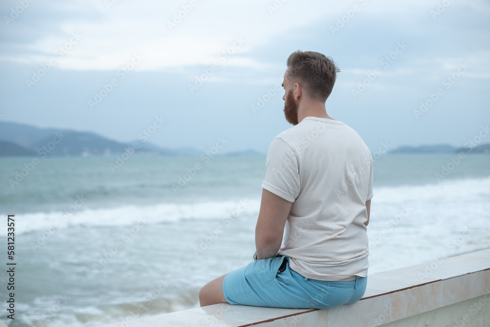 Young calm sad serious man is sitting on embankment near sea, ocean on beach, thinking, meditate on natural background Young calm sad serious man is sitting on embankment near sea, ocean on beach, thinking, meditate on natural background