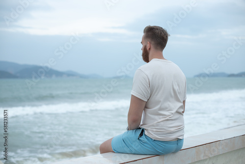 Young calm sad serious man is sitting on embankment near sea, ocean on beach, thinking, meditate on natural background 