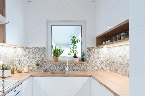 Modern kitchen with white furniture, wooden counter top, tiles pattern and sink near the window. New interior in bright apartment with stylish design.