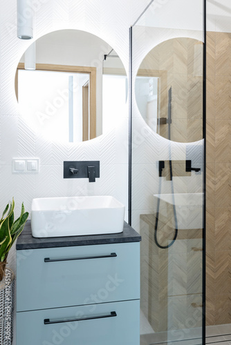 Luxurious and interior of bathroom with ceramic washbasin on the cabinet, round mirror o the white wall with led lights and shower with glass. Vertical.