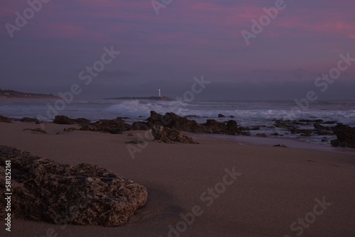 Pink hour view of the clouds above the ocean around Faro de Trafalgar lighthouse seen from Mangueta Beach, Spain, framed by rough dark gray beach rocks and wild waves rolling into Zahora Beach photo