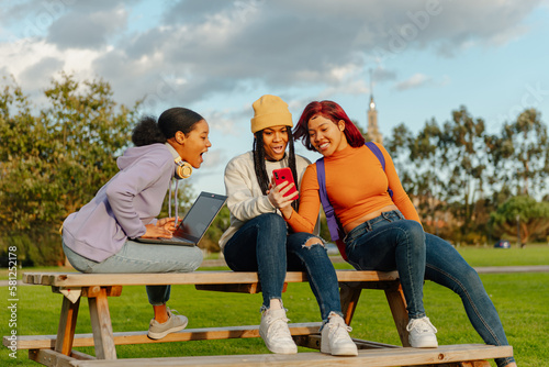 multiracial group of 3 female friends sitting on a bench on a university campus using a cell phone and a laptop. classmates having a good time during a break