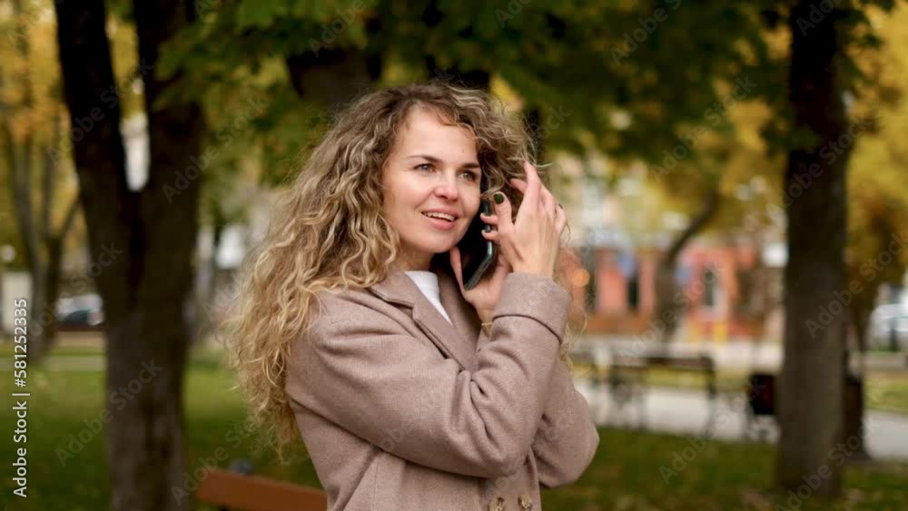 Attractive young blonde with thick wavy hair with a beautiful smile in a coat walks in the autumn park, talking on a mobile phone and smiling sweetly. Autumn Walk Attractive young blonde with thick wavy hair with a beautiful smile in a coat walks in the autumn park, talking on a mobile phone and smiling sweetly. Autumn Walk