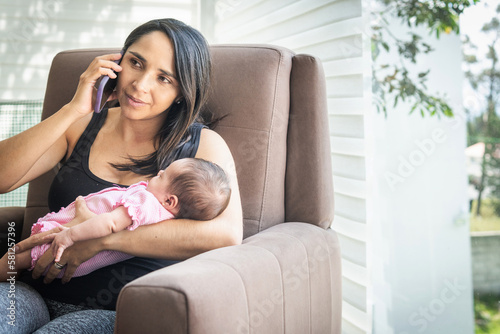 Latin woman sitting on a couch working on her cell phone with her sleeping baby at home