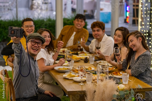 Group of Asian people using mobile phone taking selfie together during outdoor celebration dinner party in the garden on summer holiday vacation. Man and woman friend reunion meeting at restaurant.